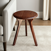 Sidney Round Side Table - Solid Walnut Wood with three splayed legs, shown in a real interior setting next to a light upholstered armchair on a textured rug.