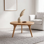 Sidney Solid Oak Wood Round Coffee Table with three tapered legs in a bright, modern living room setting next to a white armchair.