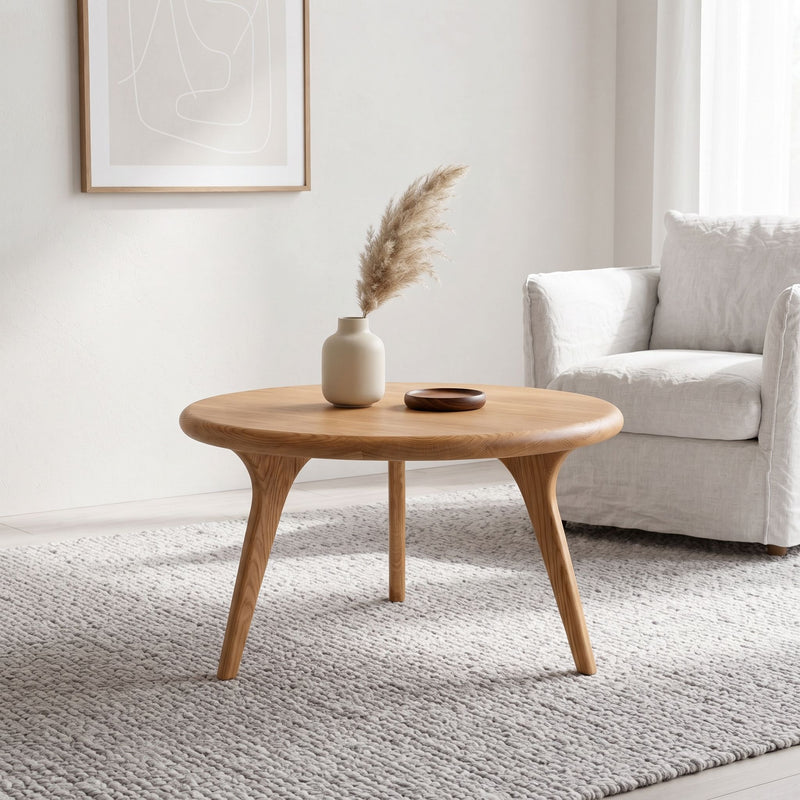 Sidney Solid Oak Wood Round Coffee Table with three tapered legs in a bright, modern living room setting next to a white armchair.