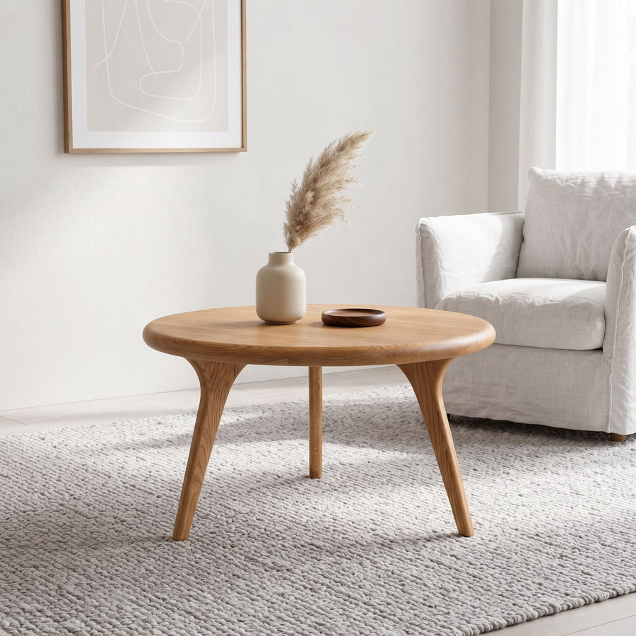 Sidney Solid Oak Wood Round Coffee Table with three tapered legs in a bright, modern living room setting next to a white armchair.
