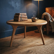 Sidney Solid Oak Wood Round Coffee Table with tapered legs in a cozy room setting, styled with a stack of vintage books and a steaming mug. A dark blue wall, a floor lamp, and a leather armchair are visible in the background.