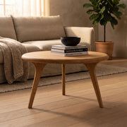 Sidney Solid Oak Wood Round Coffee Table with three tapered legs, styled in a warm living room setting with a beige sofa, books, and a black bowl on top.