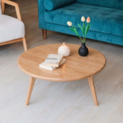 Sidney Solid Oak Wood Round Coffee Table with tapered legs in a modern living room setting, styled with a white candle, a stack of books, and a black vase with tulips. A teal velvet sofa and a white armchair are visible in the background.