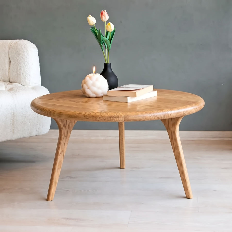 Sidney Solid Oak Wood Round Coffee Table with tapered legs, styled with a black vase of tulips, a white candle, and books on a light wood floor against a gray wall.