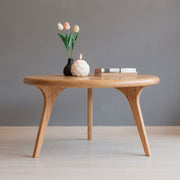 Sidney Solid Oak Wood Round Coffee Table with a rounded oval top and three tapered legs, styled with a black vase of tulips, a white candle, and a stack of books against a neutral gray background.