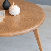 A close-up of Sidney Solid Oak Wood Round Coffee Table with a visible grain, styled with a white textured candle, a black vase, and a book on a light gray floor.