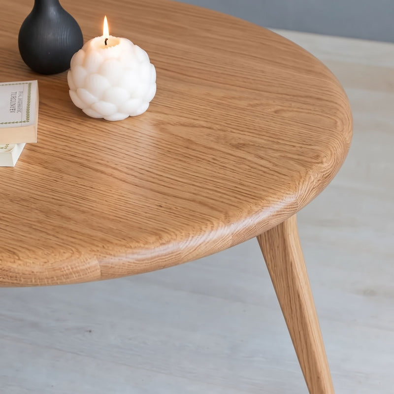 A close-up of Sidney Solid Oak Wood Round Coffee Table with a visible grain, styled with a white textured candle, a black vase, and a book on a light gray floor.