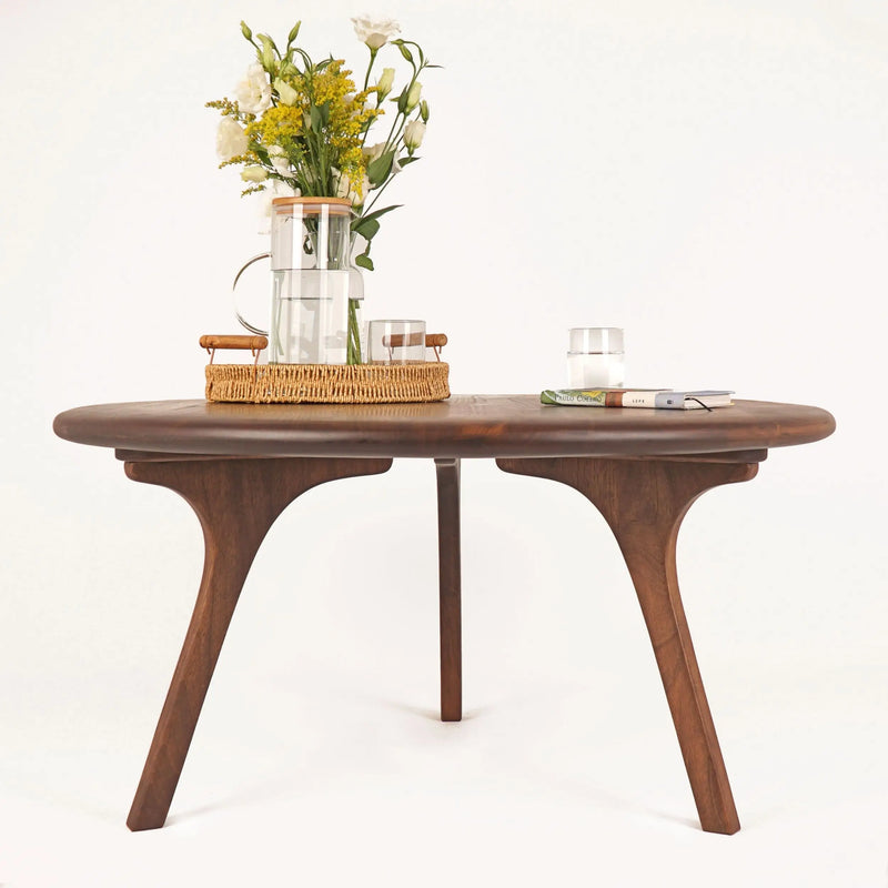 Direct front view of the Sidney Solid Wood Mid-Century Round Coffee Table on a white background, captured from a lower angle. The aesthetic, sturdy wooden legs and rich walnut wood grain are showcased, with elegant decor items arranged on the tabletop.