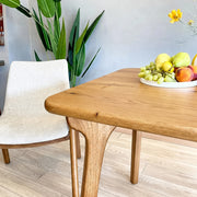 Sidney Square Dining Table Solid Oak Wood – Mid-Century Modern Design with a live edge, shown in a modern dining room setting next to a white upholstered chair. A white plate of fresh fruit sits on the table.
