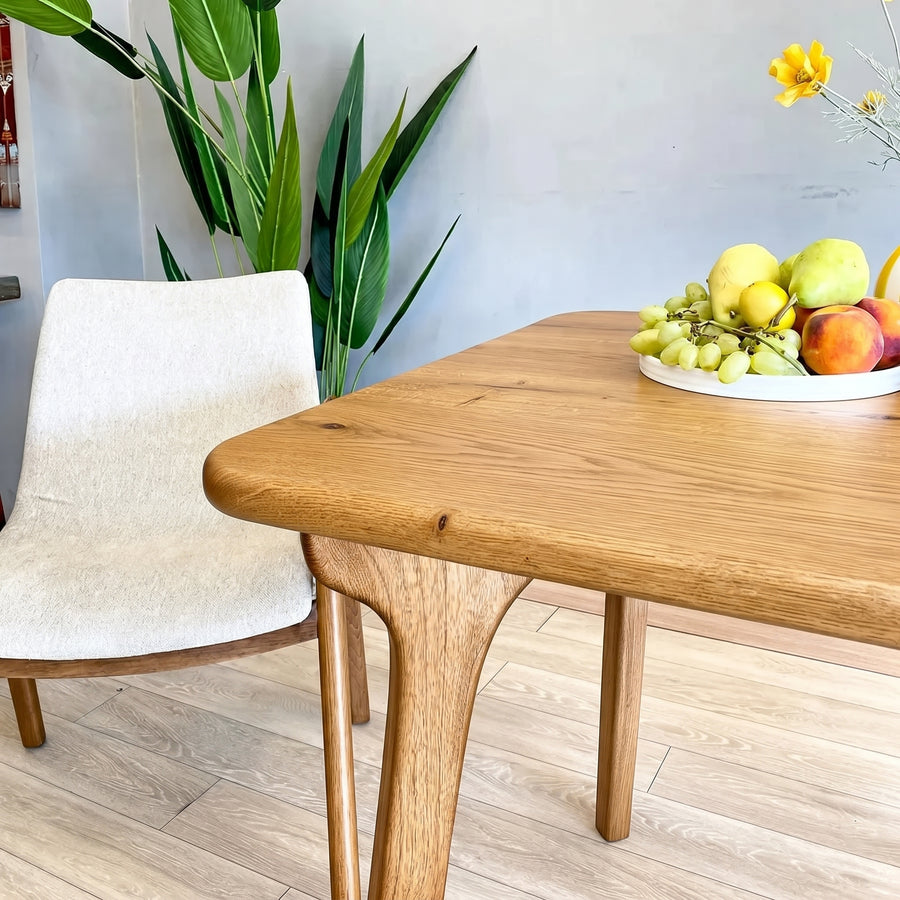 Sidney Square Dining Table Solid Oak Wood – Mid-Century Modern Design with a live edge, shown in a modern dining room setting next to a white upholstered chair. A white plate of fresh fruit sits on the table.