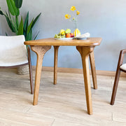 Sidney Square Dining Table Solid Oak Wood – Mid-Century Modern Design with four tapered legs and a natural oak finish, styled in a room with a gray wall, light wood floor, a green plant, and partial chairs on both sides.