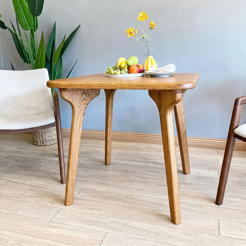 Sidney Square Dining Table Solid Oak Wood – Mid-Century Modern Design with four tapered legs and a natural oak finish, styled in a room with a gray wall, light wood floor, a green plant, and partial chairs on both sides.