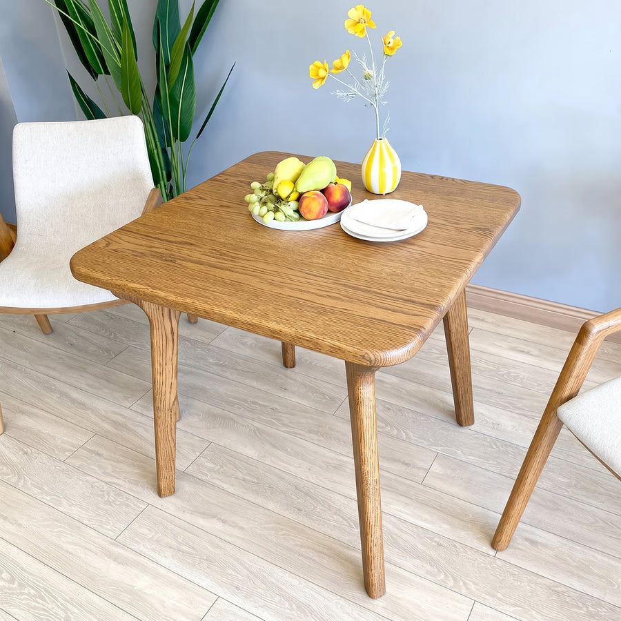 Sidney Square Dining Table Solid Oak Wood – Mid-Century Modern Design
with rounded corners and tapered legs, styled with a fruit bowl, plates, and a yellow-striped vase in a bright room with light wood flooring and chairs.