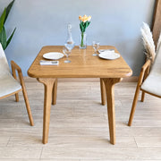 Sidney Square Dining Table Solid Oak Wood – Mid-Century Modern Design with four tapered legs, styled with plates, wine glasses, a glass decanter, and a vase of tulips, flanked by two modern chairs in a bright room.
