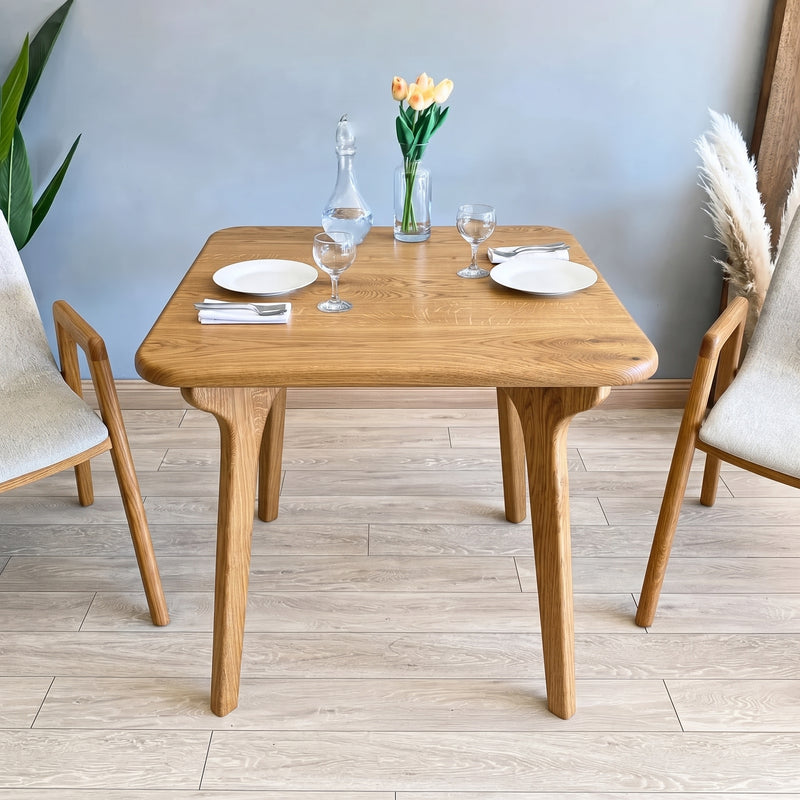 Sidney Square Dining Table Solid Oak Wood – Mid-Century Modern Design with four tapered legs, styled with plates, wine glasses, a glass decanter, and a vase of tulips, flanked by two modern chairs in a bright room.