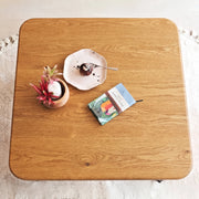 Sidney Square Wood Coffee Table Solid Oak Wood – Mid-Century Modern Design with rounded corners, styled with a small potted succulent, a plate with a chocolate dessert, and a book on a white textured rug.