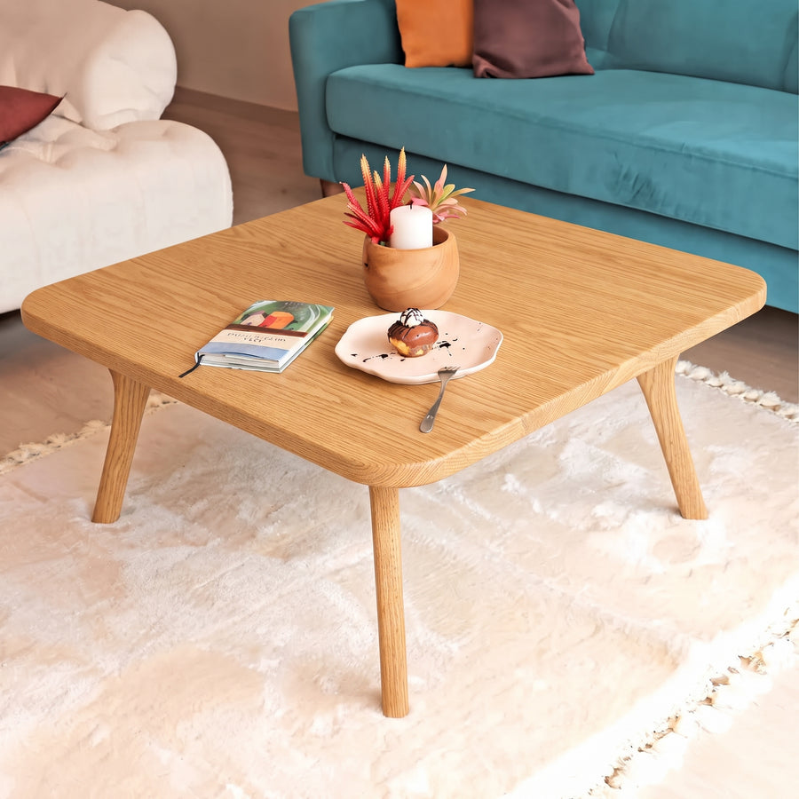 Sidney Square Wood Coffee Table Solid Oak Wood – Mid-Century Modern Design with tapered legs, styled in a cozy living room setting with a plush cream rug, a teal sofa, and decorative items on top.