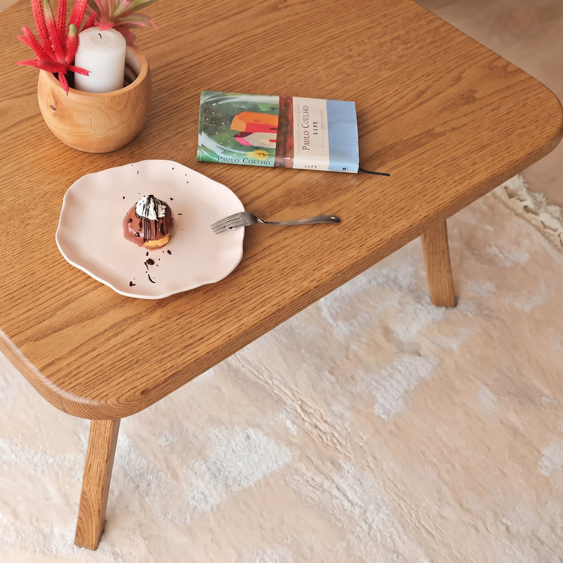 Sidney Square Wood Coffee Table Solid Oak Wood – Mid-Century Modern Design with a pink plate holding a small dessert, a fork, a book, and a small plant pot with a candle on a cream rug.