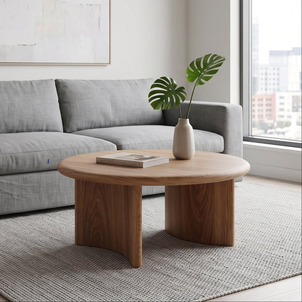 A modern living room scene featuring Soren Round Oak Coffee Table | Sculptural Scandinavian Modern Design in the foreground, a light gray fabric sofa behind it, a neutral rug, and a large abstract framed print on the wall.