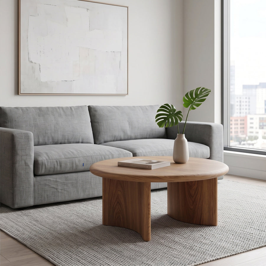 A modern living room scene featuring Soren Round Oak Coffee Table | Sculptural Scandinavian Modern Design in the foreground, a light gray fabric sofa behind it, a neutral rug, and a large abstract framed print on the wall.