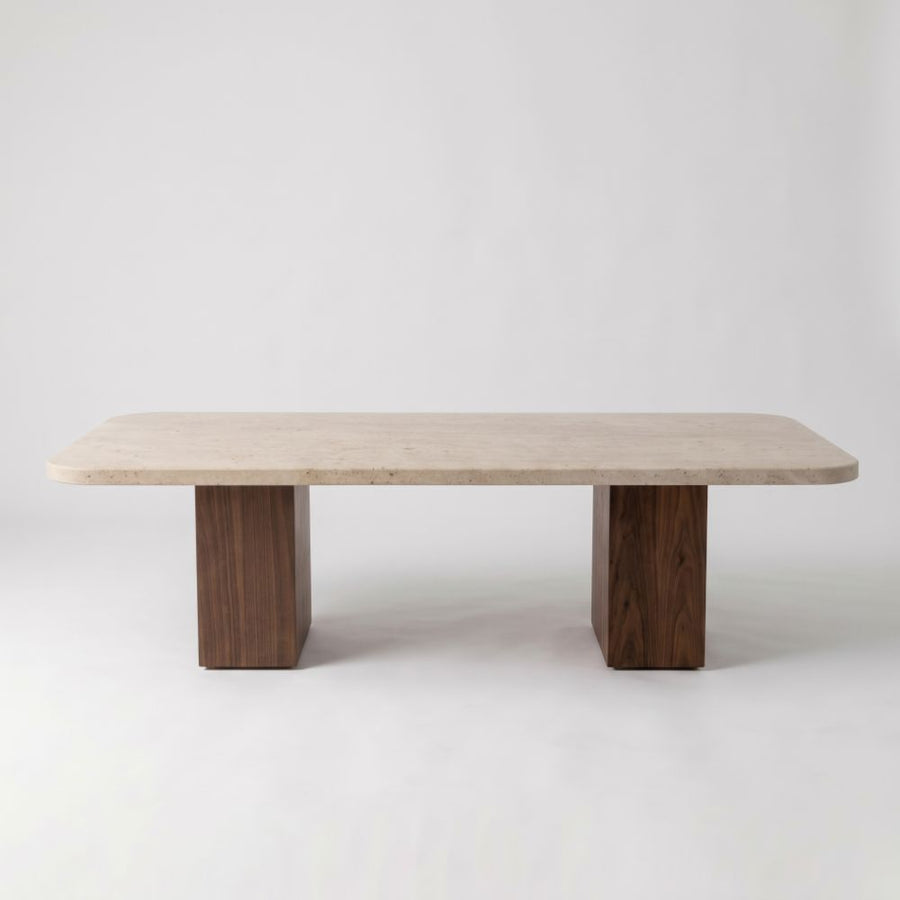 Eye-level straight-on studio shot of Wilder Rectangular Travertine Coffee Table featuring a rounded-corner beige stone top and dual solid walnut column bases