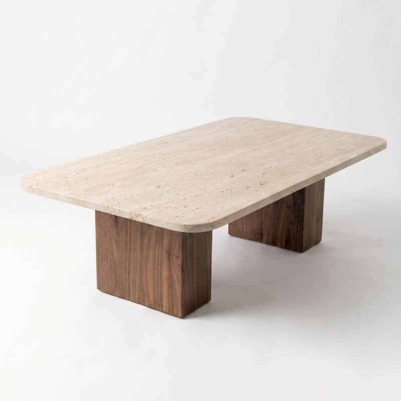 Three-quarter studio view of Wilder Rectangular Travertine Coffee Table featuring a rounded-corner beige stone top and dual solid walnut column bases