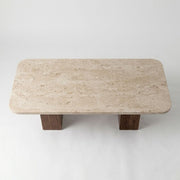 Detailed studio shot from high angle showing rounded-corner beige travertine stone top surface and dual solid walnut column bases on Wilder Rectangular Coffee Table