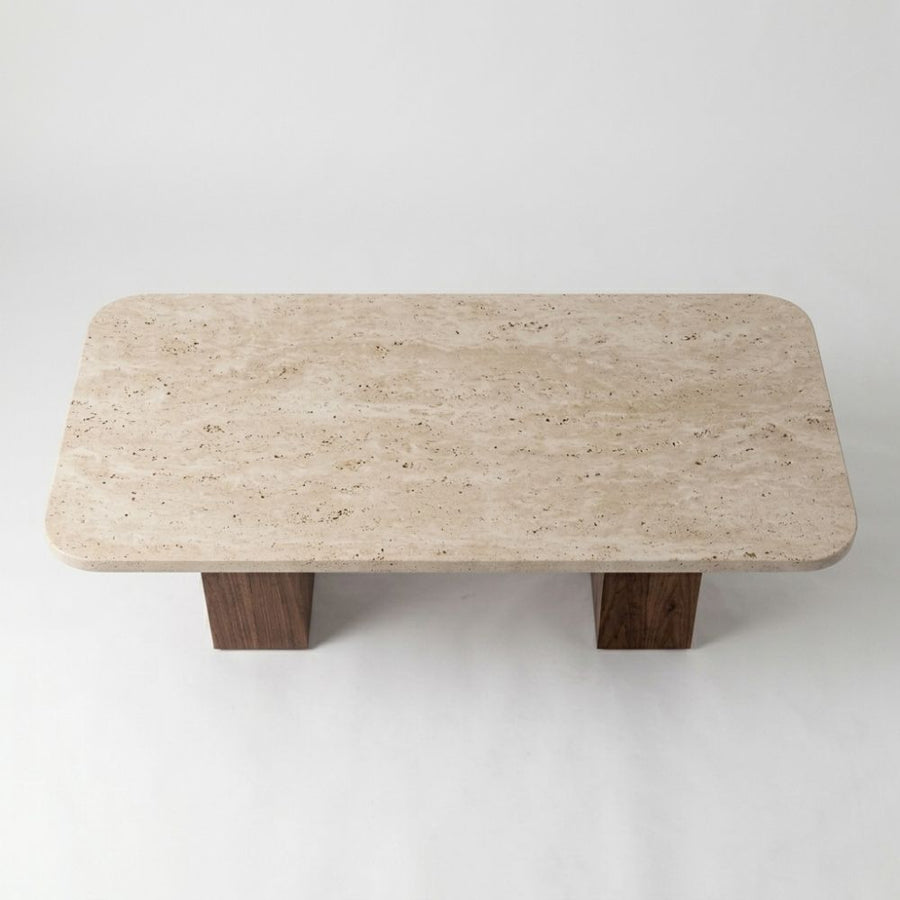 Detailed studio shot from high angle showing rounded-corner beige travertine stone top surface and dual solid walnut column bases on Wilder Rectangular Coffee Table