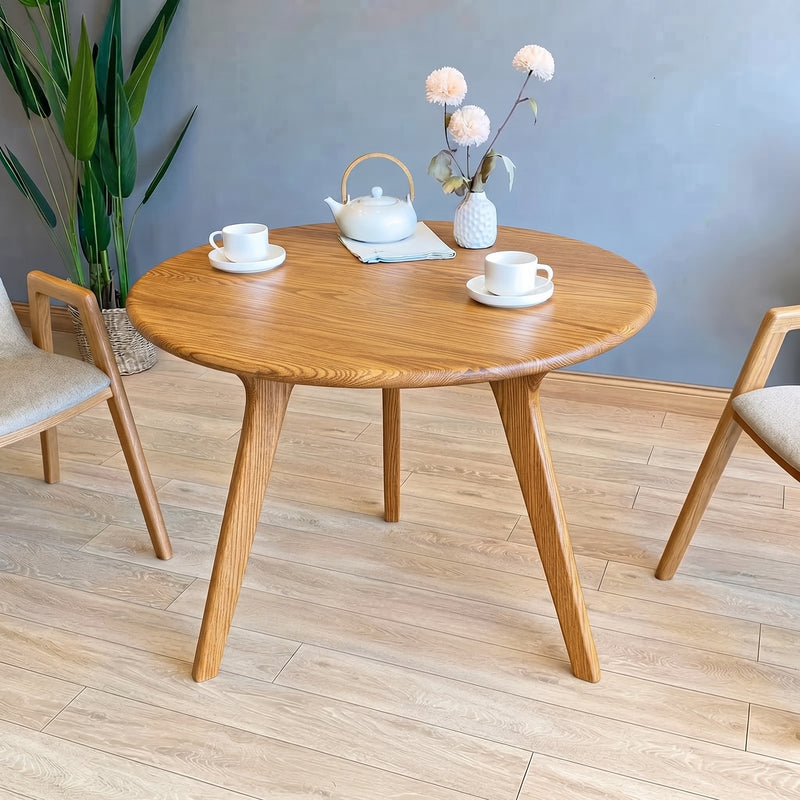 Sidney Circular Dining Table Solid Oak Wood – Mid-Century Modern Design with tapered legs, styled with a white teapot, two teacups, and a vase of flowers, set in a modern room with a gray wall and light wood flooring.